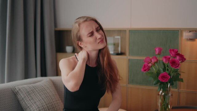 Young woman on a couch massaging her sore neck, wincing from tension and pain caused by stress, fatigue or injury while seeking relief, experiencing neck pain at home