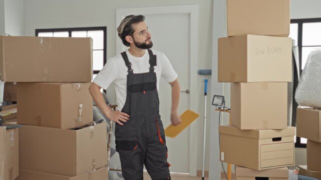 Man with hand on hip surveying stacked cardboard boxes and holding a package in a building; thoughtful moving preparation.