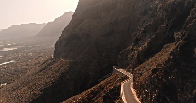 Winding mountain road carved into steep, rugged cliffs of Tenerife near Masca Valley, distant cultivation fields under a bright sky, adventure and journey. Dramatic landscape. Aerial view drone flight
