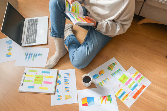 Person sitting on floor with laptop and coffee, reviewing colorful charts, graphs, and a marketing strategy mind map on paper, indicating flexible remote work and data analysis