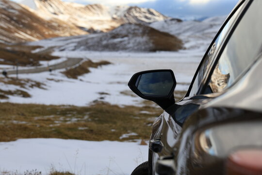 Snow-dusted mountains rise behind a parked car. Side mirror reflects the vast, quiet landscape. Driver's hand rests on the gearshift, hinting at pause or anticipation
