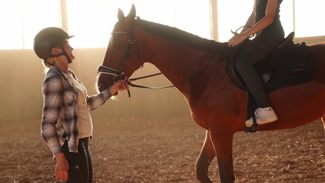 Female instructor teaching young girl to ride bay horse in indoor arena at sunset. Equestrian school for children, professional horse training lesson in golden hour light, sport, education, mentorship