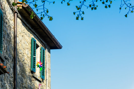 View of a traditional stone building with green shutters and a colorful pinwheel toy against a clear blue sky in Loro Ciuffenna, Tuscany, Italy.