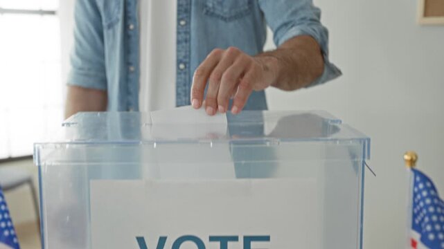 Man drops ballot into clear ballot box beside small us flags on table in a building; patriotic participation.