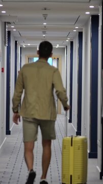 Solo Man With Luggage. Person Proceeding With Suitcase In Contemporary Hotel Hallway. Single Traveler Steadily Navigating Modern Hotel Corridor Towards Exit With Yellow Rolling Bag