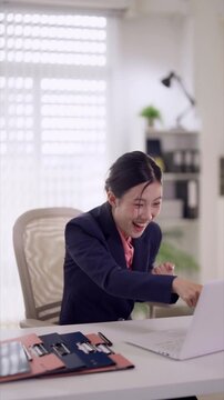 excited Asian businesswoman celebrating success achievement raise fists winner gesture sitting desk laptop computer modern office expressing joy excitement