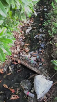 A heavily polluted narrow ditch filled with plastic waste, debris, and stagnant water, surrounded by green foliage, highlighting environmental degradation.