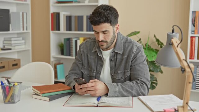 Man holds smartphone and scrolls while seated at desk with books, open notebook and lamp in a building study space; concentration study.