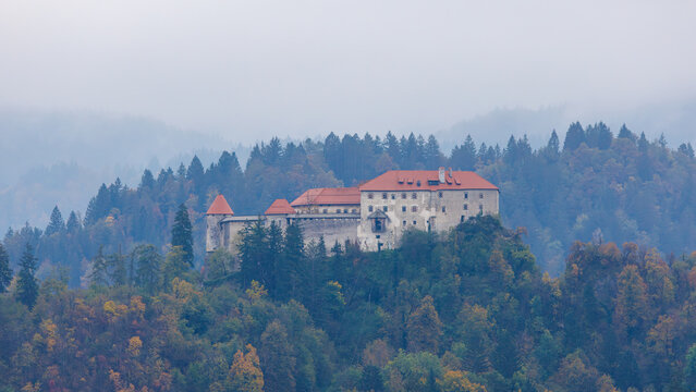 View of Bled Castle perched on a cliff surrounded by dense evergreen and autumn-colored forests under a misty sky in Bled, Radovljica, Slovenia.