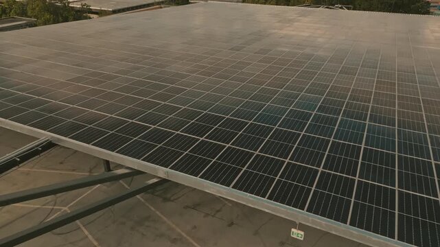 Dense solar grid generating renewable electricity stretching across wide parkade roof toward distant ocean waters bordered by coastal mountains beneath cloudy sky captured by low skimming aerial motio