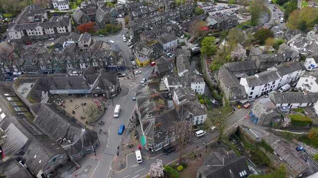 A forward drone shot flies over the bustling northern streets of Ambleside, Cumbria, revealing the market square, busy roads, stone buildings, and spring blossom trees below.