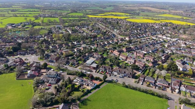A left orbital drone shot over Goffs Oak, Hertfordshire, revealing suburban streets, green gardens, and vibrant yellow rapeseed fields stretching into the English countryside beyond.