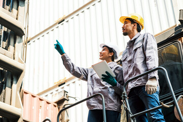Fototapeta premium Two industrial workers in helmets inspect shipping containers at a freight yard; one points while the other holds a tablet during a safety check.