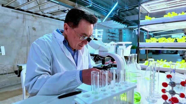 Scientist in a modern lab uses a microscope beside test tubes, flasks, and molecular models, while LED grow lights illuminate plants on shelves in the background.