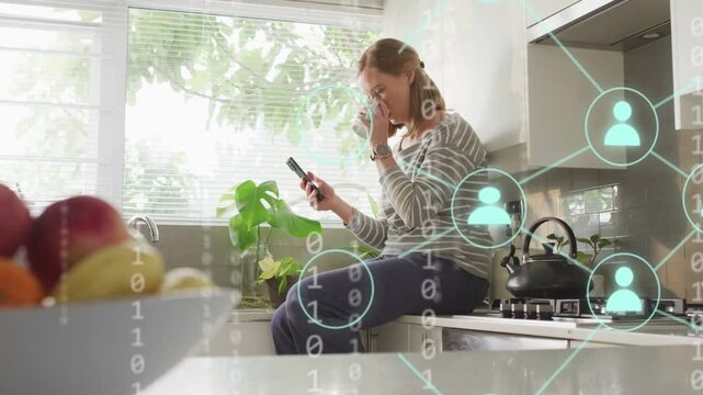 Woman sitting on counter tapping phone launching binary or network icons showing tech data then fadi