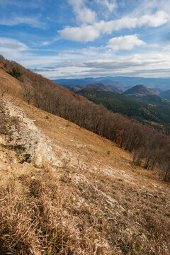 View of Kozi chrbat mountain ridge with a steep rocky slope, leafless autumn trees, and rolling forested hills under a cloudy blue sky in Mostenica, Banska Bystrica Regio, Slovakia.