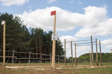 Fototapeta premium Wooden post with number 2 sign and red flag on an outdoor track. White powder marks the path on the grass. Forest and blue sky background