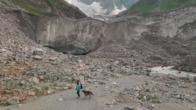 Aerial view woman Hiker cuddle stray dog by Shkhara glacier in Svaneti region. Massive snow-capped Mount Shkhara peaks dominate horizon. Upper Svaneti alpine landscape near Ushguli, Georgia.