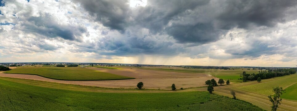 Panoramiczny widok na malownicze pola uprawne pod dramatycznym, zachmurzonym niebem. Nad horyzontem widoczne lokalne opady deszczu. Wiejski krajobraz rolniczy w letni dzień przed burzą.
