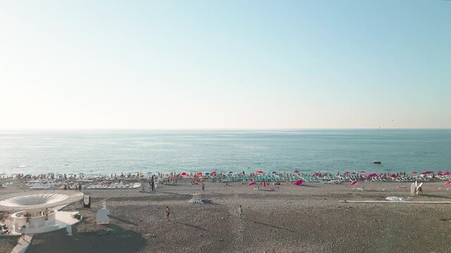 Low angle aerial panoramic fly over view of busy pebble beach in Batumi, Georgia, featuring rows of pink umbrellas, sunbathers, and calm Black Sea waters on clear summer evening at sunset