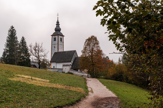 View of the historic Church of St. John the Baptist with its tall bell tower and stone walls surrounded by autumn trees and a grassy hill in Municipality of Bohinj, Radovljica, Slovenia.