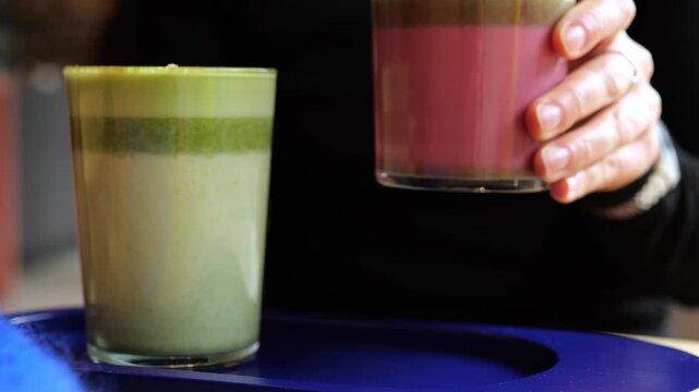 Close up of a barista's hand serving two colorful, layered superfood lattes in glasses on a blue tray at a coffee shop, showcasing a healthy green matcha tea and a pink beetroot beverage