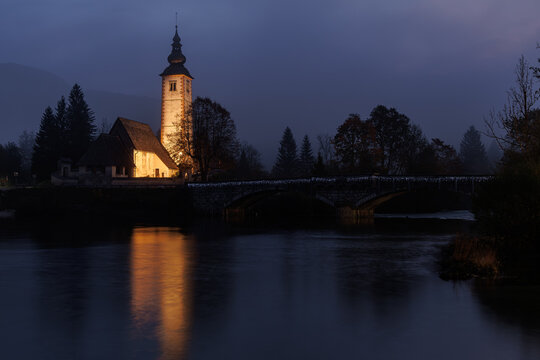 View of the illuminated Church of St. John the Baptist and the stone bridge reflected in the calm waters of Lake Bohinj at night Municipality of Bohinj, Radovljica, Slovenia.