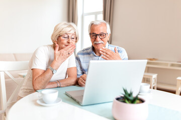Fototapeta premium Happy senior couple blowing air kisses to laptop screen during video call. Elderly man and woman using computer for remote communication at home. Retirement technology and family connection concept.