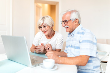 Fototapeta premium Cheerful elderly couple watching grandchildren on laptop screen with joy. Senior man and woman having video chat with family at dining table. Concept of technology, aging, and family connection.