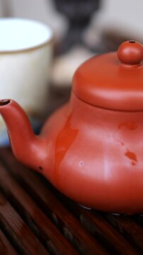 Close up of a red yixing clay teapot sitting on a wooden tea tray during a traditional chinese tea ceremony, with hot water dripping down its side and a small white cup in the background