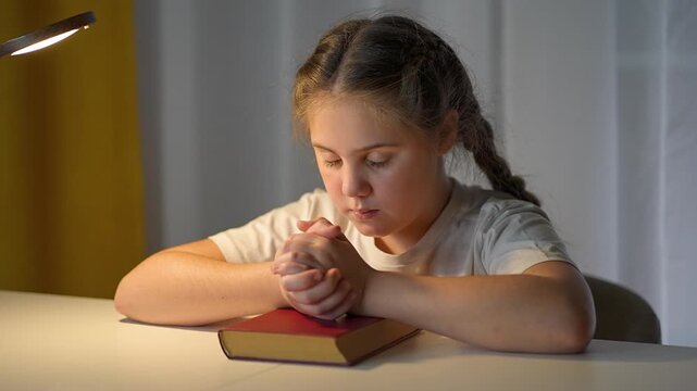 Child folds hands in steady prayer with closed eyes, resting quietly on book. Girl focuses in worship peaceful devotion. Still moment of belief shows harmony between child, peace, and deeper belief
