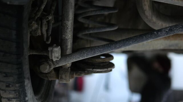 Close up of a dirty car's rear suspension, including the coil spring and tire, with a mechanic performing diagnostic and maintenance work in the blurred background of a professional garage