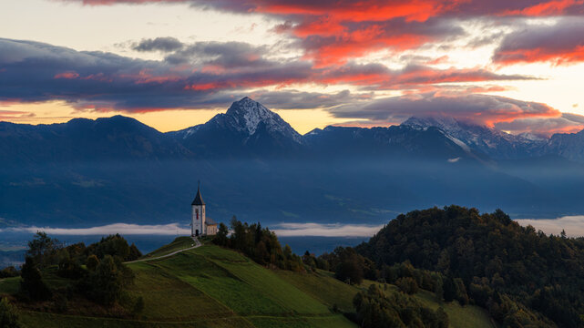 View of the Church of St. Primus and Felician on a hilltop with the Kamnik-Savinja Alps under a dramatic sunset sky in Jamnik, Kranj, Slovenia.