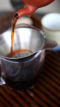 Close up of black tea pouring from a traditional clay teapot through a metal strainer into a glass pitcher during a classic chinese gongfu cha ceremony, showcasing the rich color and aroma
