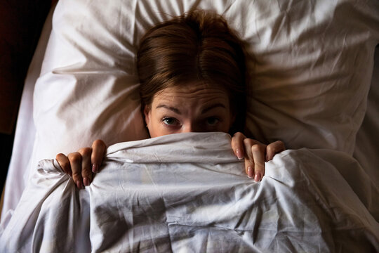 A woman lies in her bed, pulling the white blanket up to her eyes. She peeks out, eyes wide, looking at something with possible fear in the dark of the bedroom at night.