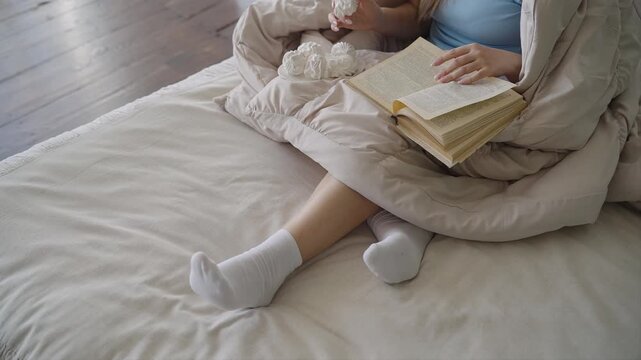 Woman in white socks sitting on bed under blanket and reading book and eating marshmallows