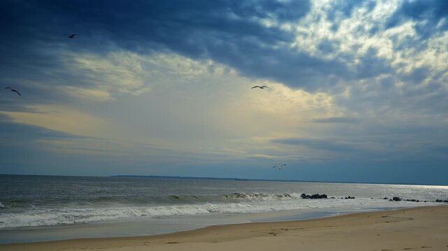 Dramatic sky over breaking waves on Atlantic coast. Breaking waves on a sandy beach with birds flying under a dark cloudy sky and a cargo ship in the distance.