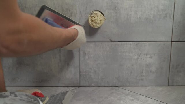 Close up of worker applying gray grout to new ceramic tiles on wall. Tiler uses grout float to fill joints between tiles during home renovation or construction project. Detailed manual labor footage