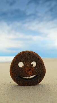 Smiley face cleaning sponge standing on a sandy beach. Circular cleaning sponge with a smiley face cut-out sitting on the sand with blurred ocean background.