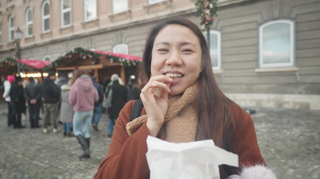 A woman enjoys a delicious snack at an outdoor market, surrounded by festive stalls and visitors. The atmosphere is lively and inviting, with decorations enhancing the experience.