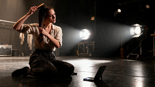 [Young woman] [with calm mood] [brushing her hair in a dimly lit studio]