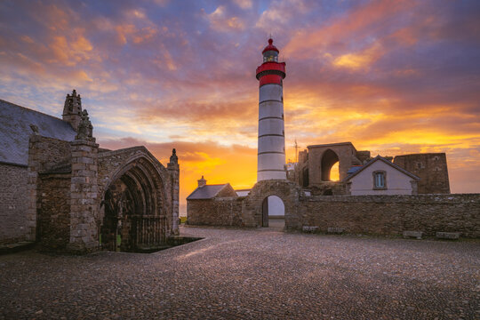 View of the Pointe Saint-Mathieu lighthouse and the ruins of the ancient abbey under a dramatic sunset sky in Plougonvelin, Brittany, France.