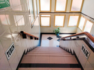 Fototapeta premium Quiet tiled descending staircase with wooden handrails, up (naik) and down (turun) signs, clean floor tiles, a potted plant, soft lighting through frosted windows, for classic vintage interior ideas.