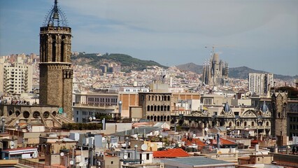 Obraz premium Barcelona cityscape with the neo-gothic cathedral bell tower and sagrada família under construction, historic rooftops and distant mountains on a clear sunny day
