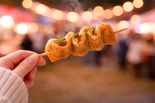 Steaming eomuk (fish cake) skewer seasoned with green onions and chili flakes being held at a vibrant night festival.