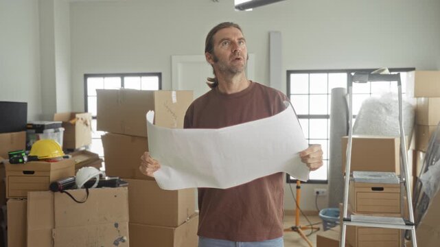 Man holds blueprint with both hands and studies plans amid stacked boxes and a ladder in building; concentration planning.