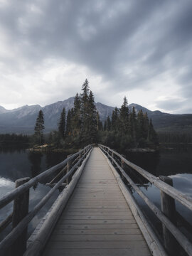 View of a wooden walkway leading to a small island with tall pine trees on a calm lake with mountains under a cloudy sky in Canada.