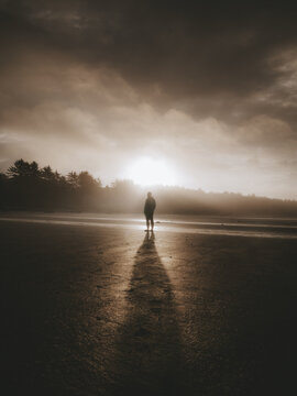 View of a person standing on a sandy beach under a dramatic cloudy sky at sunset in Canada.