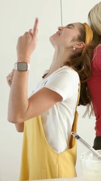 Vertical video: Singing woman in yellow overalls miming with wooden spoon in kitchen, mixing bowl