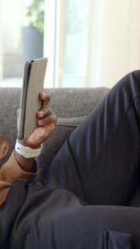 Vertical video: Lifting grey folio tablet, man tapping and swiping on couch with white smartwatch